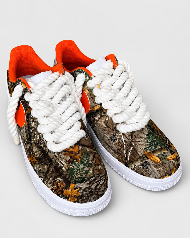 Woodland camo sneakers with white rope laces and orange lining on a light gray studio background